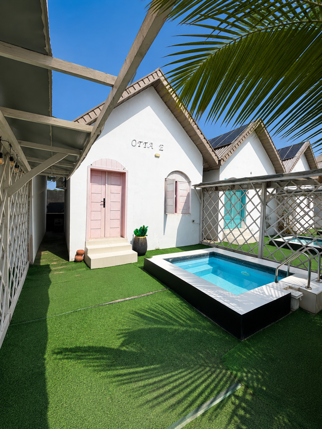 A white gabled cottage with a pink door beside a private plunge pool and green lawn